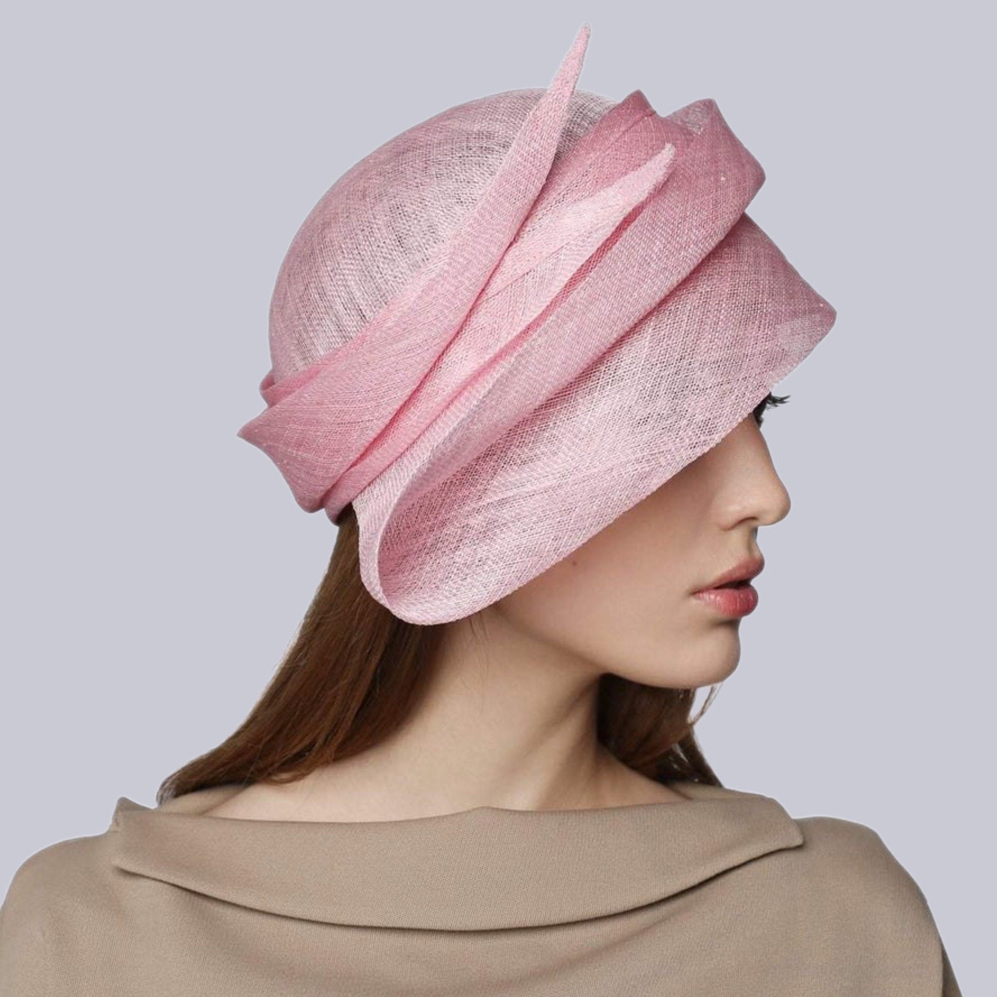 A light pink hat with a layered design. The hat features a rounded crown and asymmetrical brim with a textured weave.