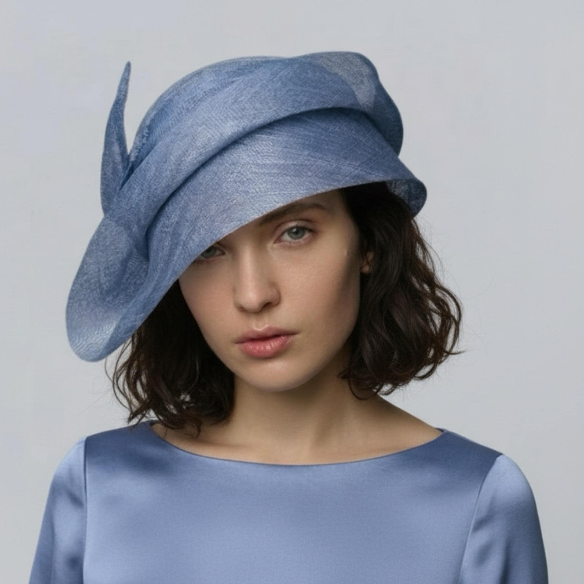 Elegant woman wearing a sculptural blue sinamay straw couture hat with a sweeping wide brim for wedding guest style.